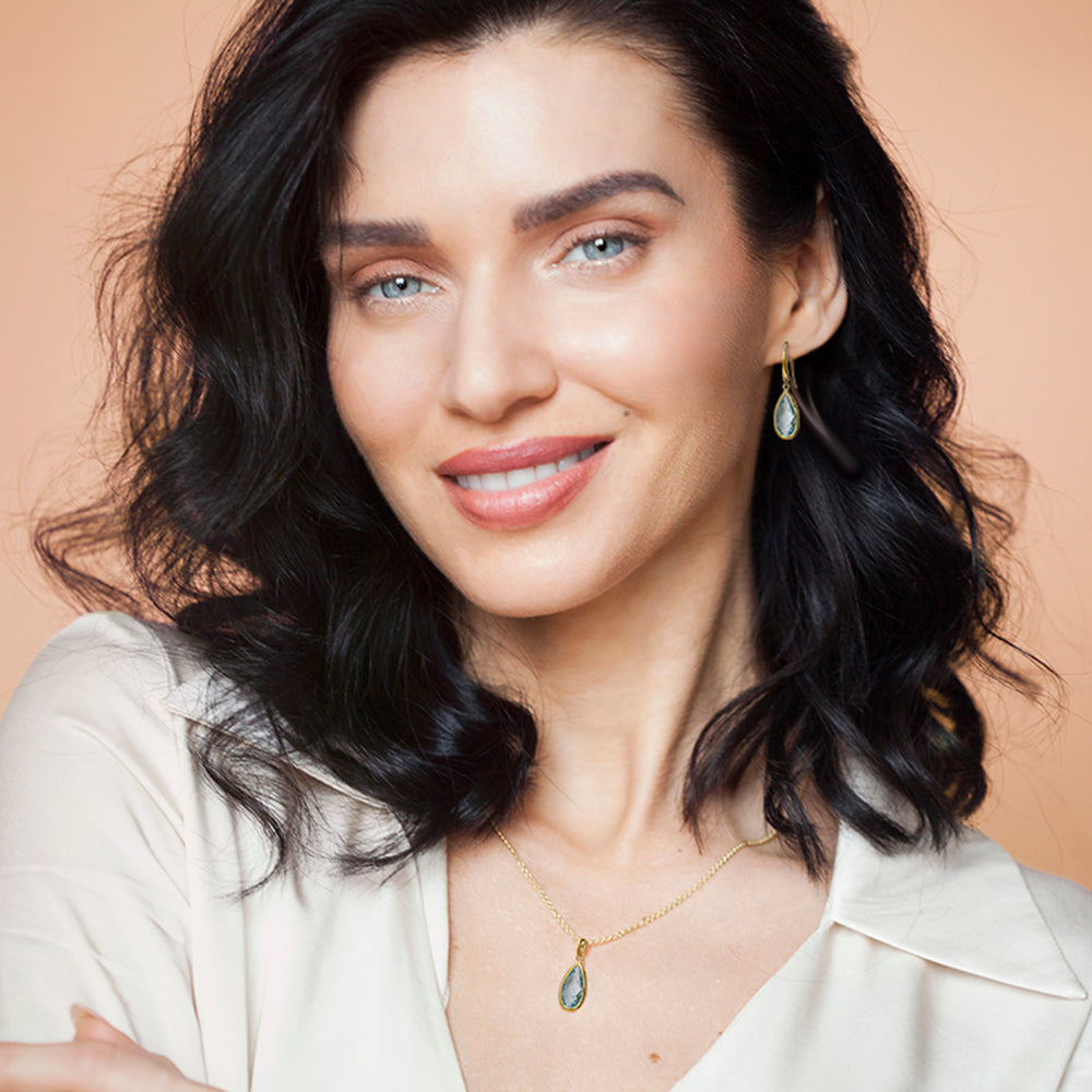 A close-up portrait of a woman wearing blue topaz pear drop earring and necklace set with gold vermeil