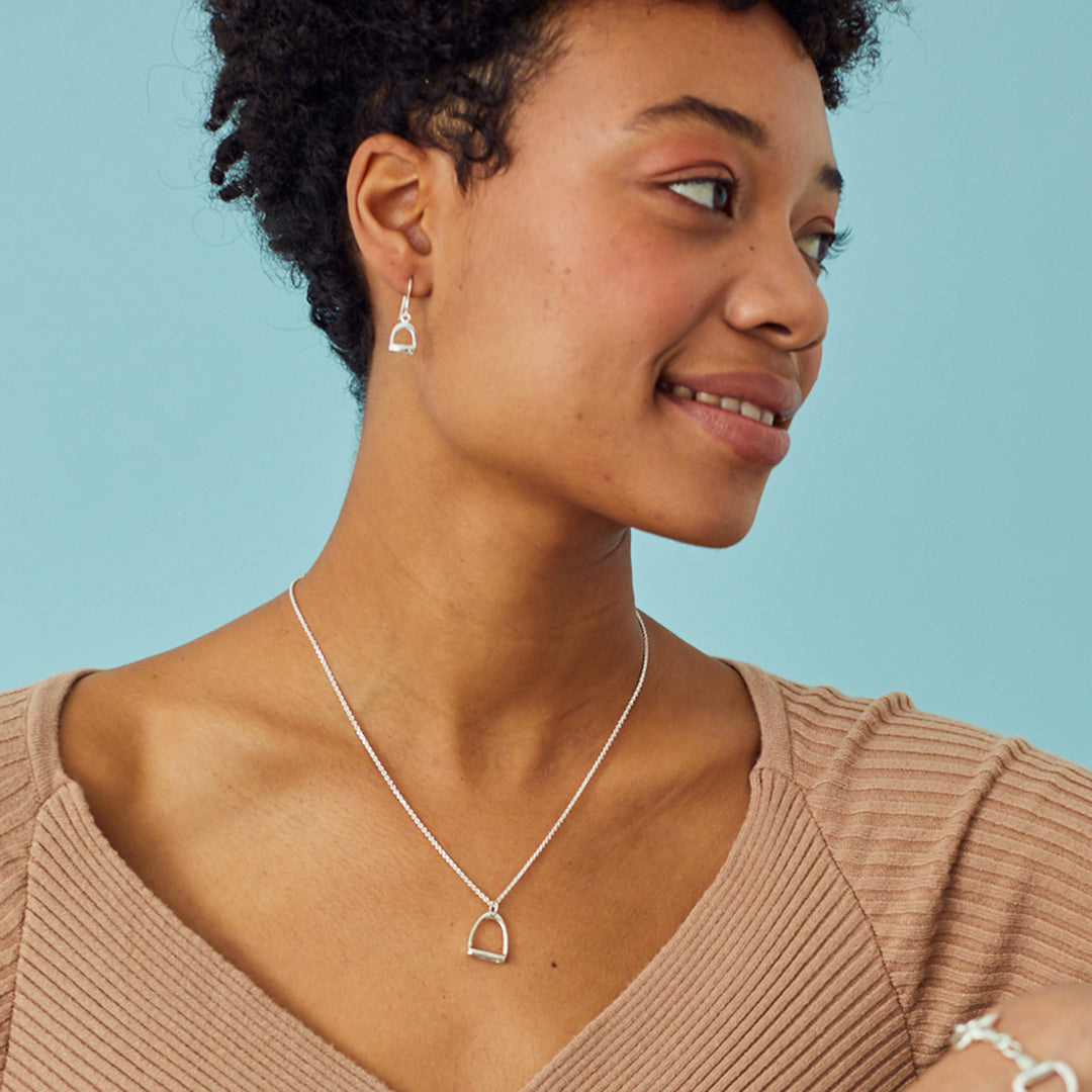 Woman wearing a silver stirrup necklace and matching earrings against a blue background