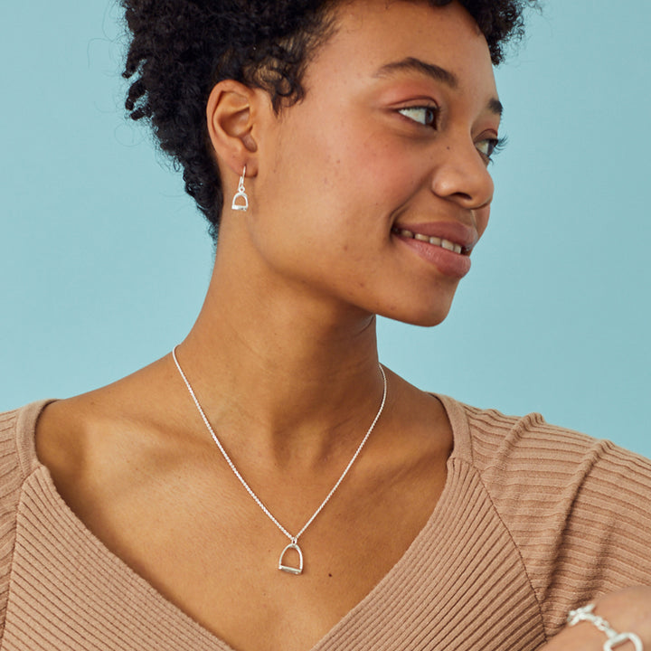 Woman wearing a silver stirrup necklace and matching earrings against a blue background