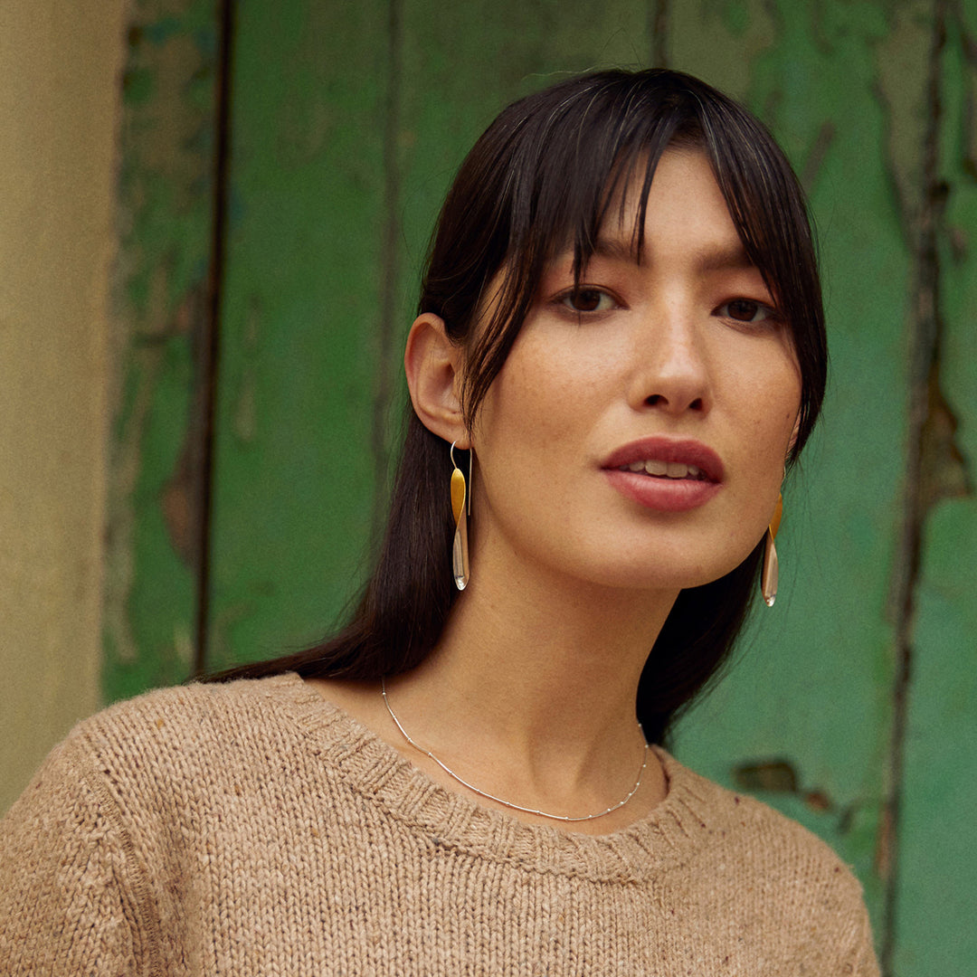 Woman wearing silver and gold dangle earrings and a beige sweater with a green door in the background