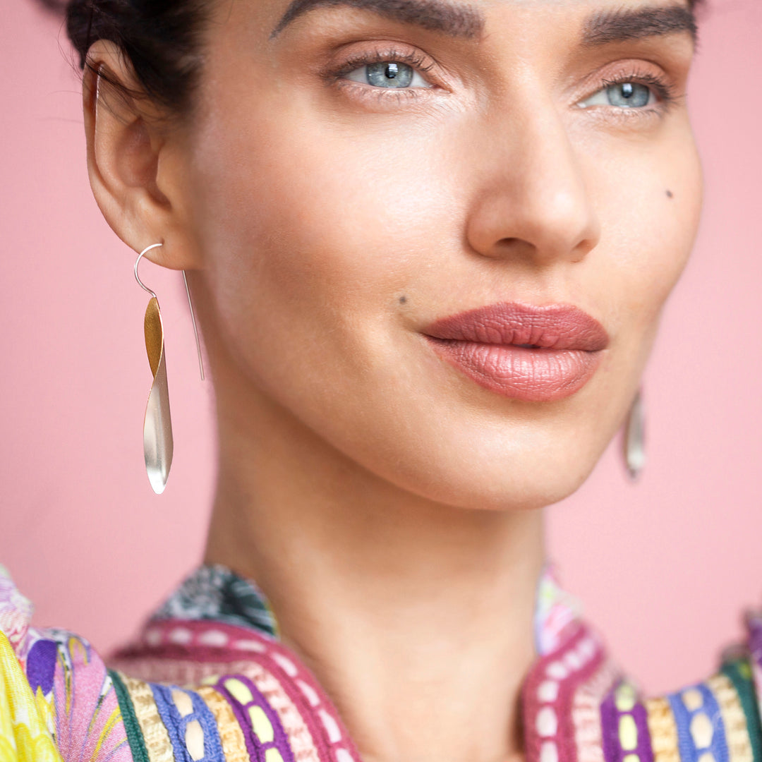 Close-up of a woman wearing long two-tone silver and gold earrings with a pink background