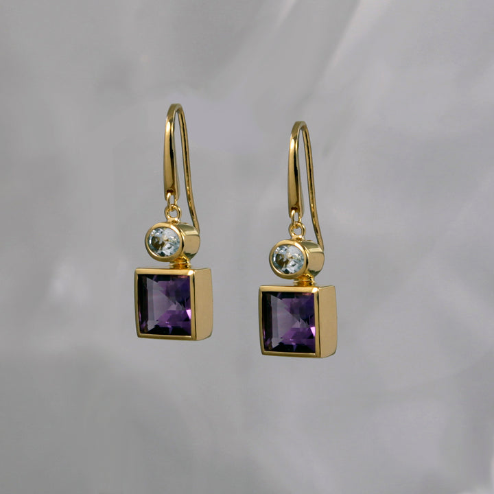 Square gold earrings with purple and light blue gemstones on a gray background