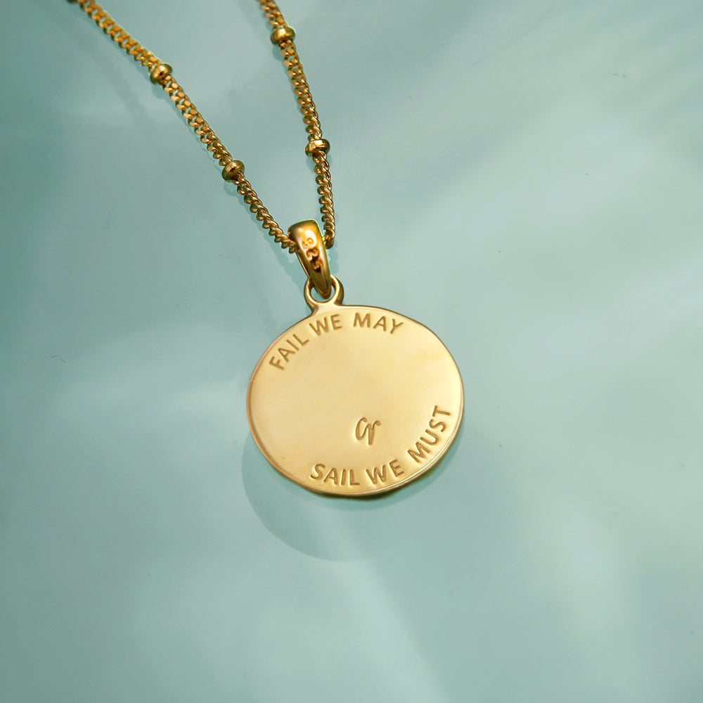 Reverse side of a gold anchor necklace with a engraved message on an aqua background