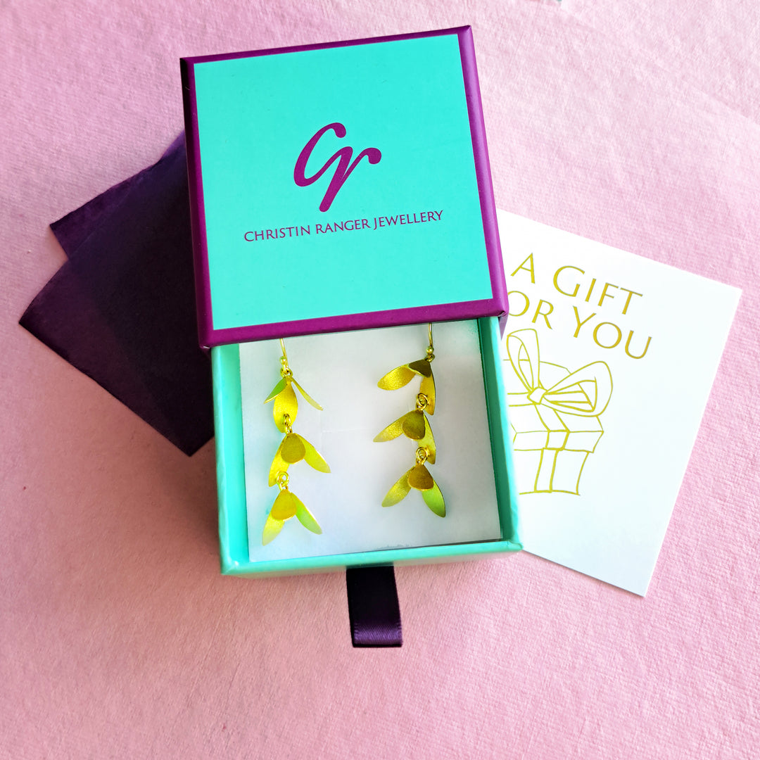 A pair of gold leaf drop earrings displayed in an open turquoise gift box with a purple lid