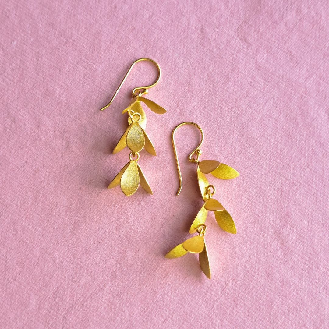 A pair of gold dangle earrings with leaf-like layered charms, displayed on a textured pink background.