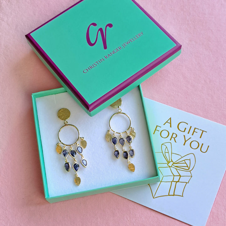 Gold and blue gemstone chandelier earrings in a Christin Ranger Jewellery gift box and card on a pink background.