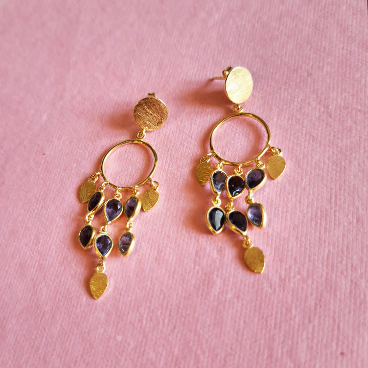 Gold chandeiier style earrings with teadrop shaped purple iolite gemstones  on a pink background.