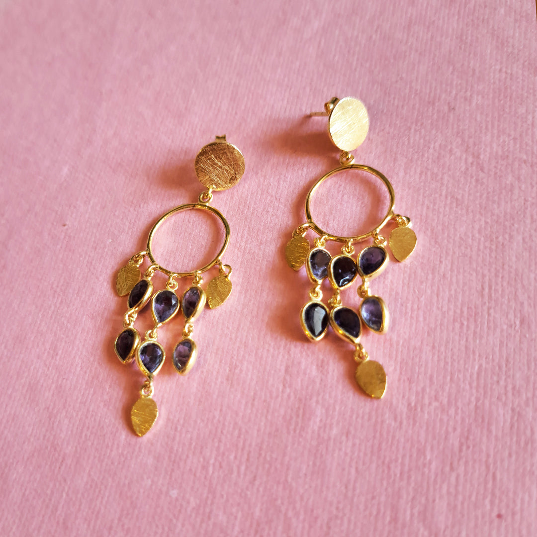 Gold chandeiier style earrings with teadrop shaped purple iolite gemstones  on a pink background.