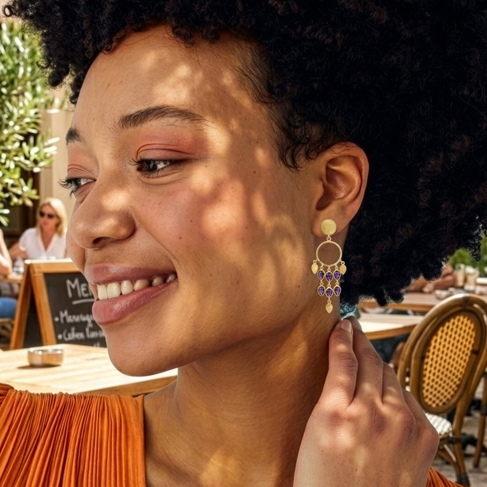 Woman wearing gold chandelier earrings with teardrop shaped iolite gemstones in a sunny plaza.