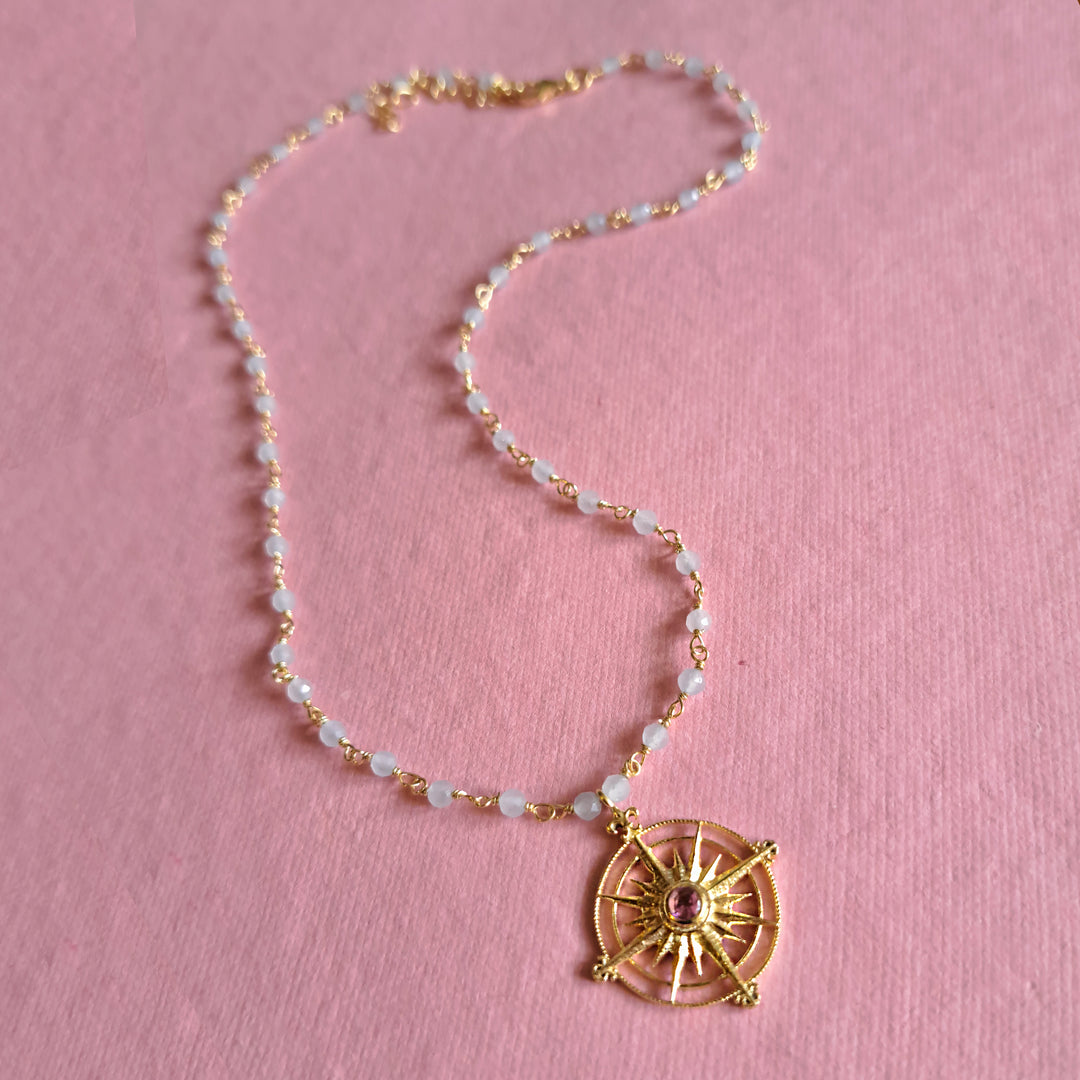 Gold sun medalion with a tourmaline gemstone in the center on a blue chalcedony beaded necklace on a pink background.