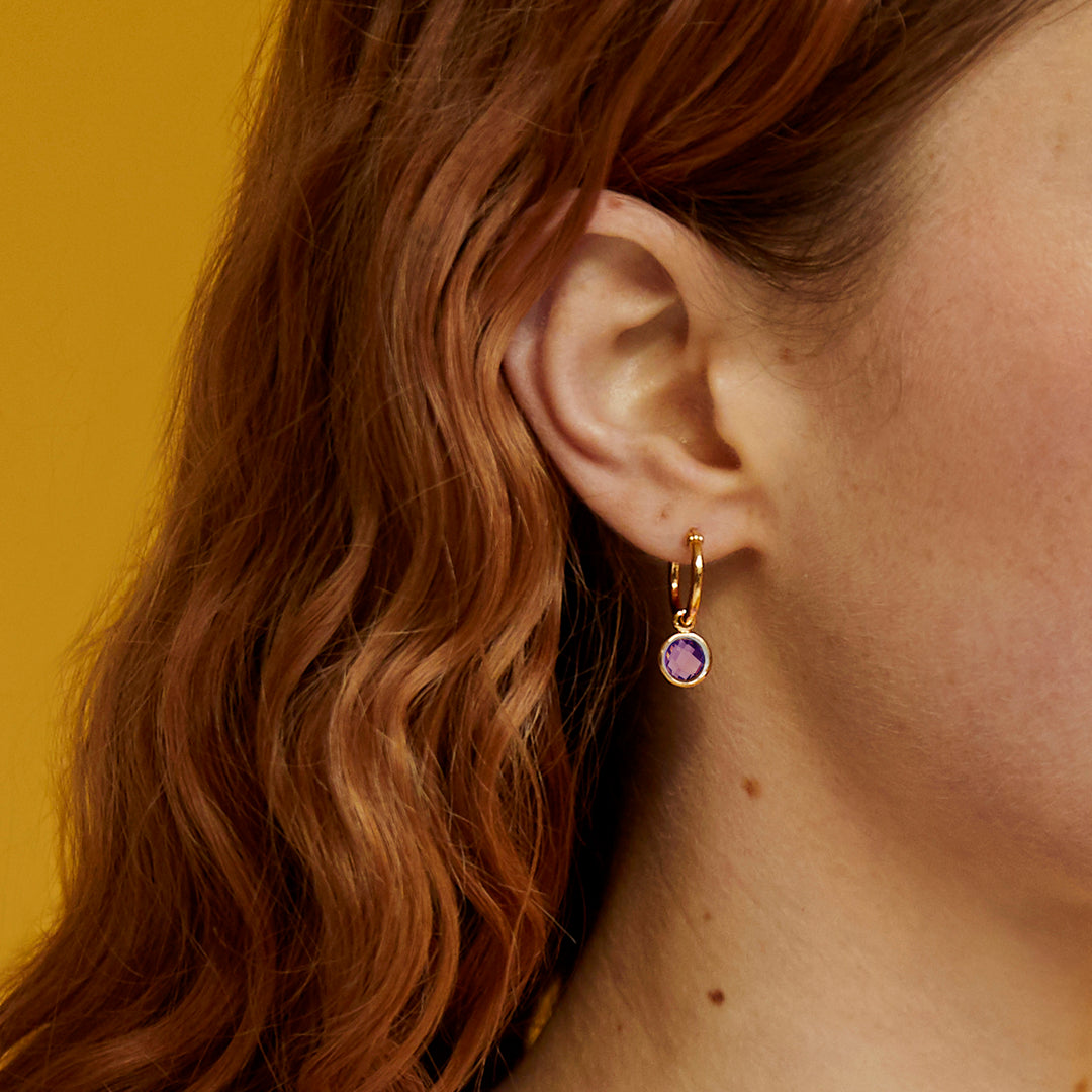Close-up of an model wearing a gold hoop earring with a purple amethyst against a yellow background