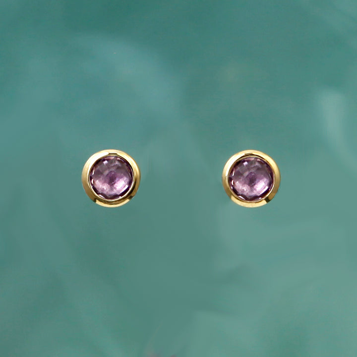 Round gold post earrings with purple gemstones on an aqua background
