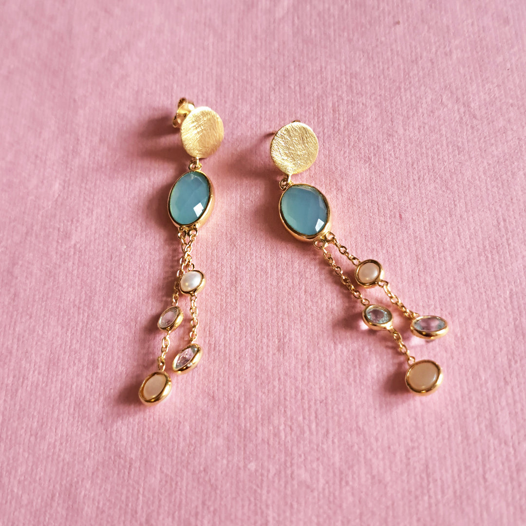 Gold drop earrings with large chalcedony stones and  chain dangles with blue topaz and pearls on a pink background.