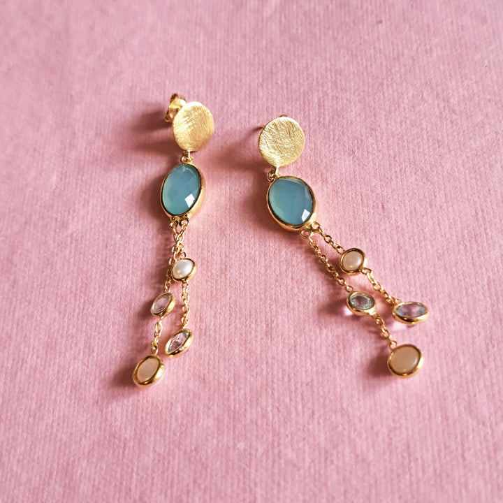 Gold drop earrings with large chalcedony stones and  chain dangles with blue topaz and pearls on a pink background.