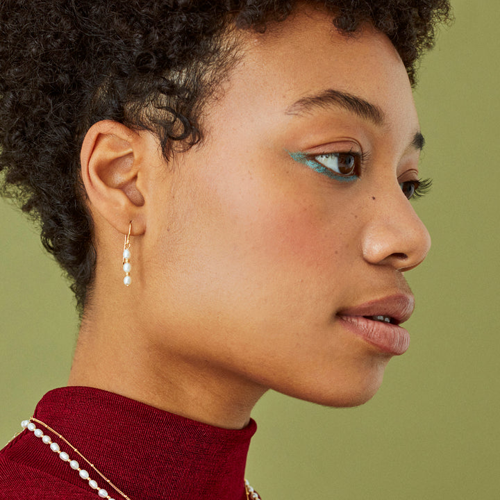 Side view of model wearing a dangle pearl and gold earring with a green background