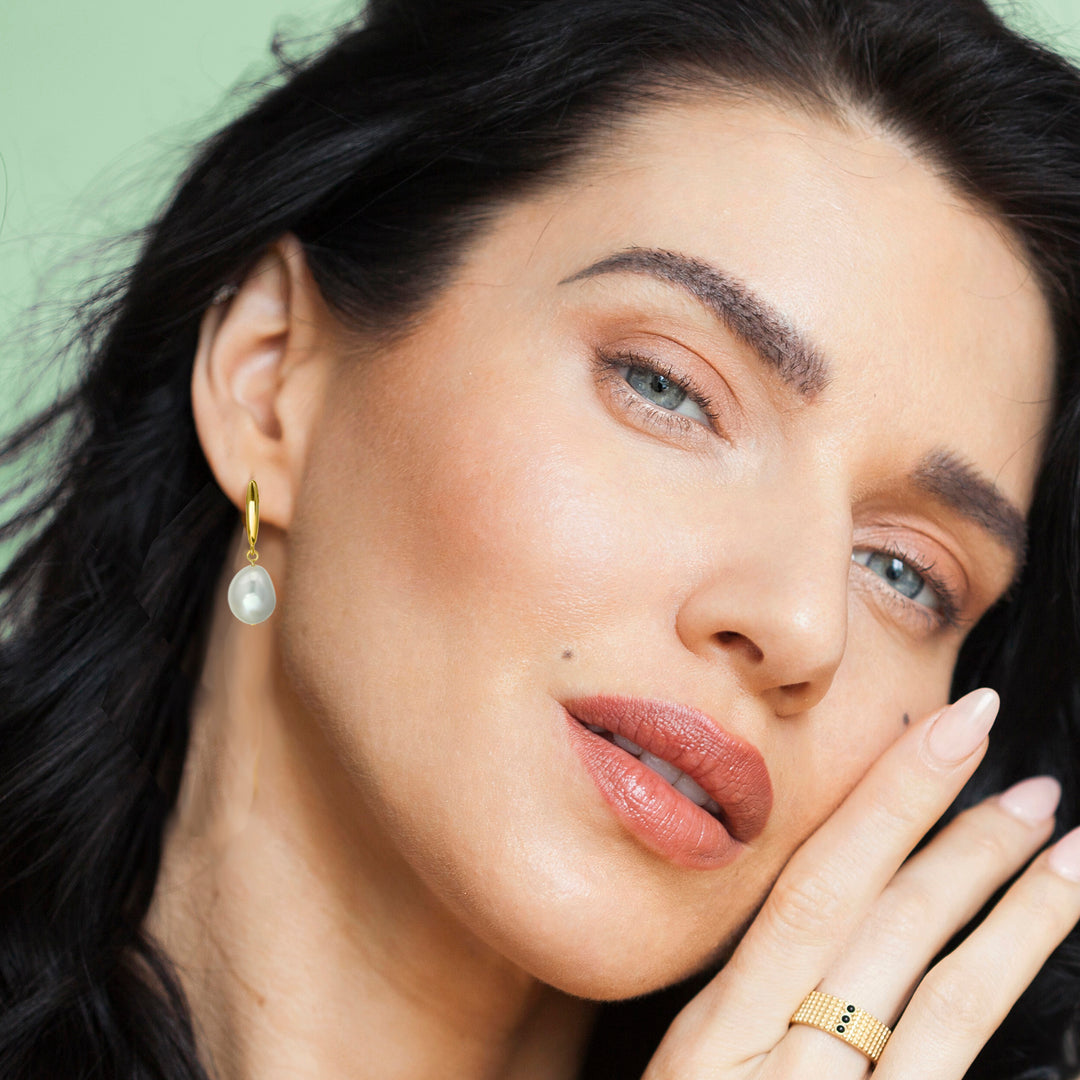 Close-up of a woman with dark hair wearing pearl earrings and a gold ring against a green background