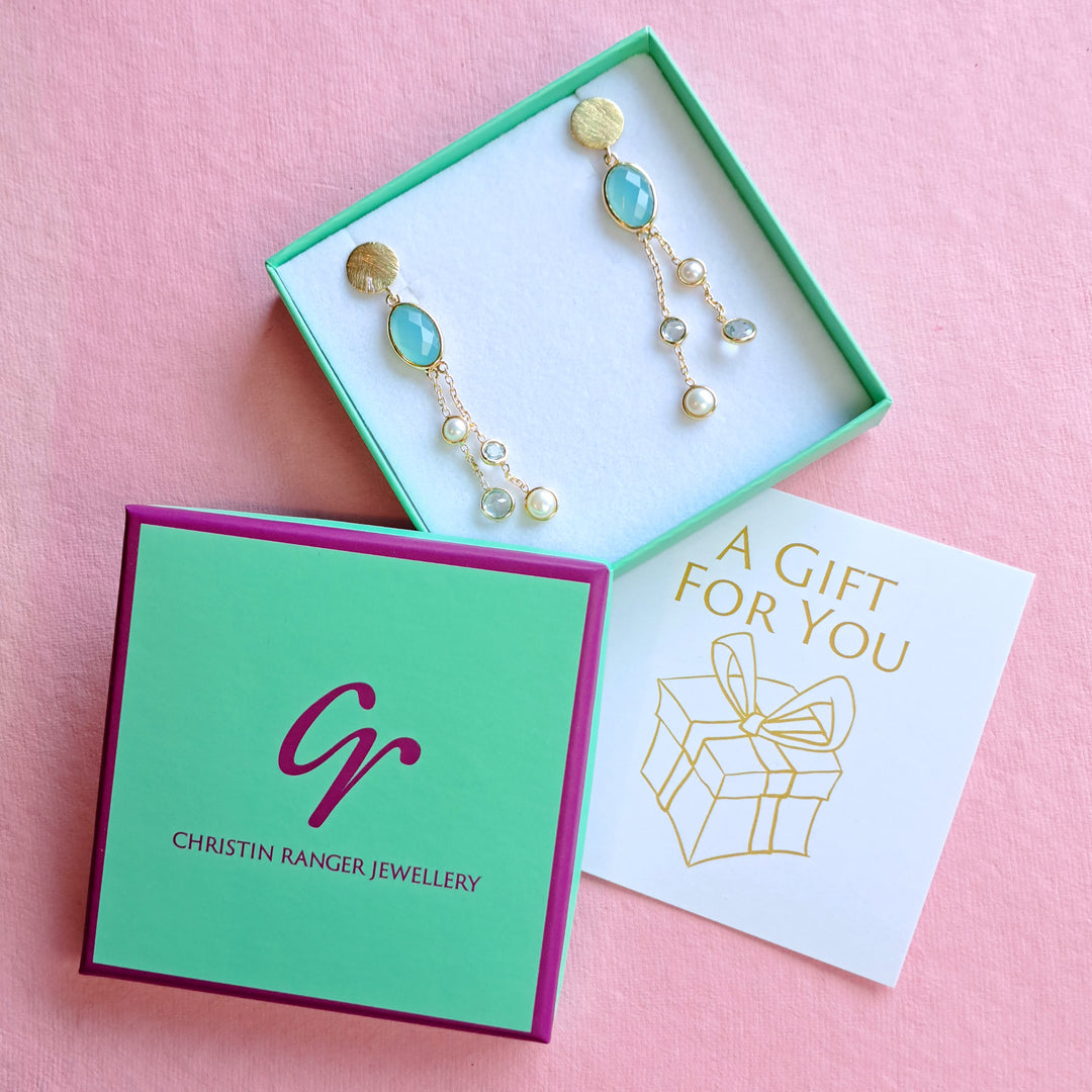 Gold statement earrings with blue gemstones and freshwater pearls in a Christin Ranger gift box  on a pink background.