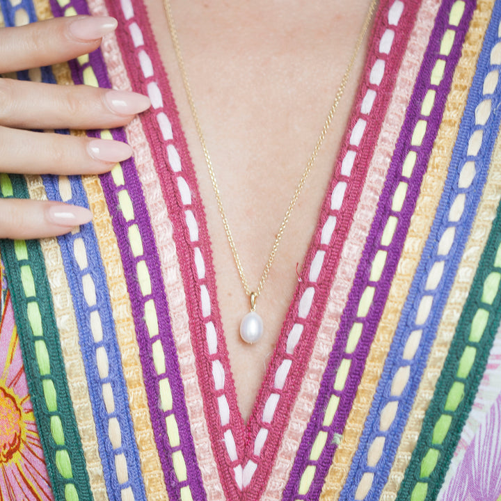 Person wearing a gold necklace with a pearl pendant against a colorful striped garment.