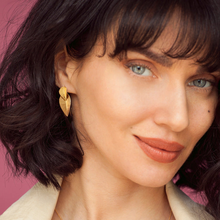 Close-up of a woman wearing gold leaf-shaped earrings with a pink background