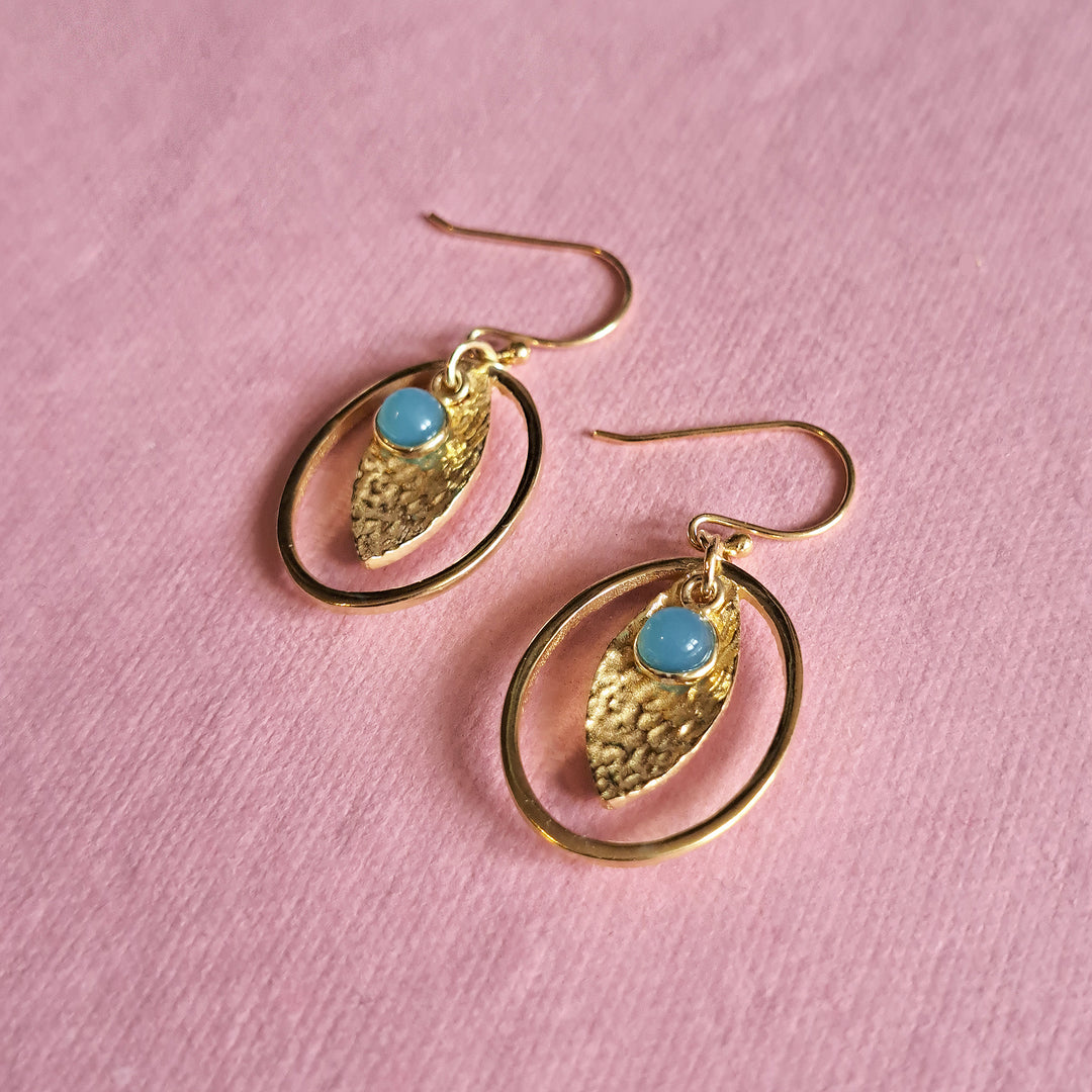 Gold oval hoop earrings with textured drops and blue chalcedony cabochons on a pink background.