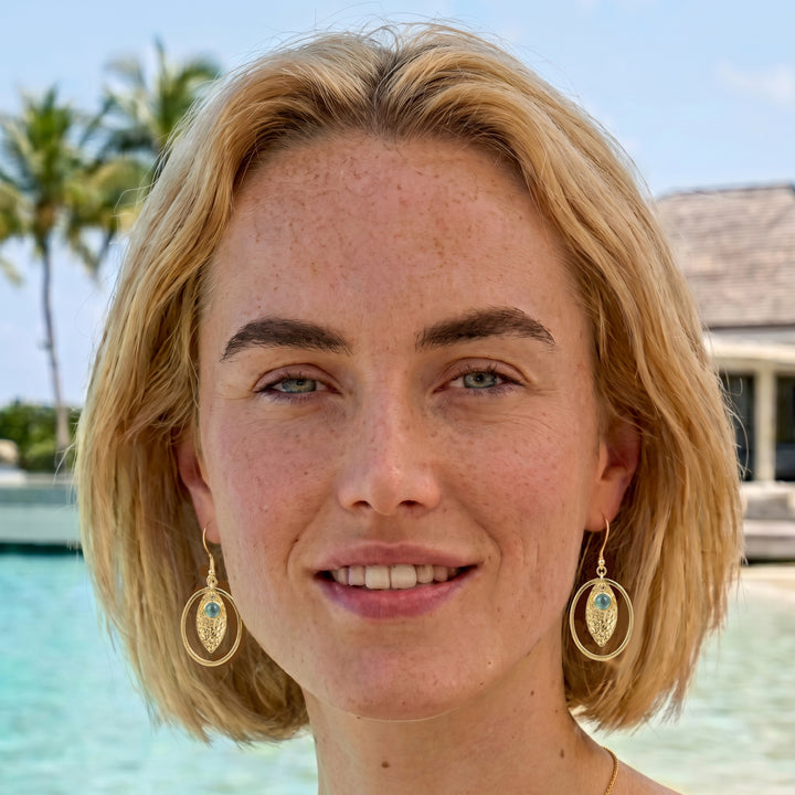 Model wearing gold textured hoop drops with blue chalcedony cabochons at the beach.