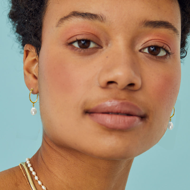 Close-up of a woman wearing  gold hoop earrings with pearls against a light blue background