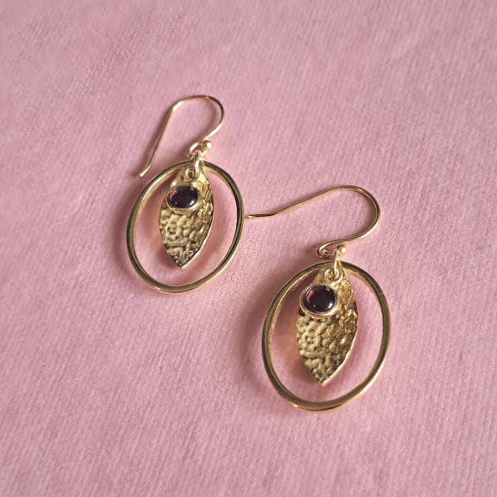 Gold hoop earrings with garnet cabochon drops on a pink background.