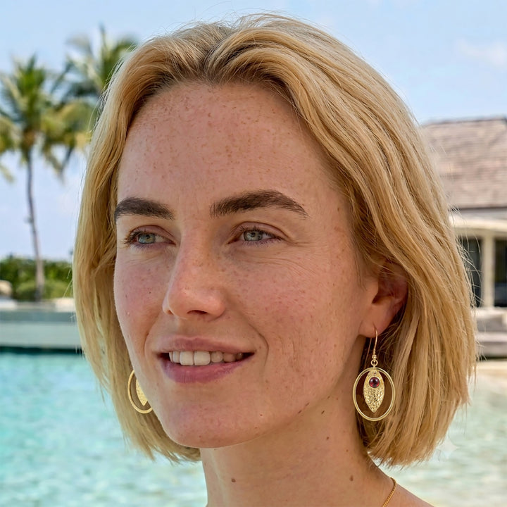 Woman wearing gold textured drop earrings with oval hoops and garnet cabochons at the beach.