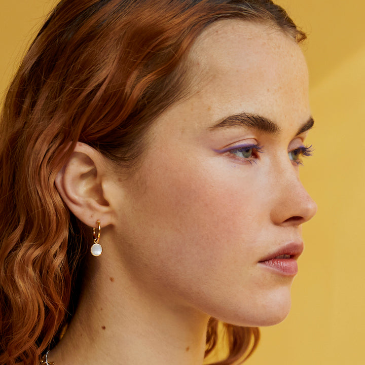 Model wearing gold huggie style hoop earrings with moonstone drops on a yellow background.