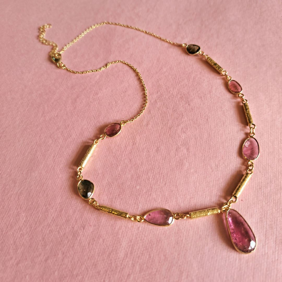 Gold necklace with green and pink tourmaline gemstones on a pink background.