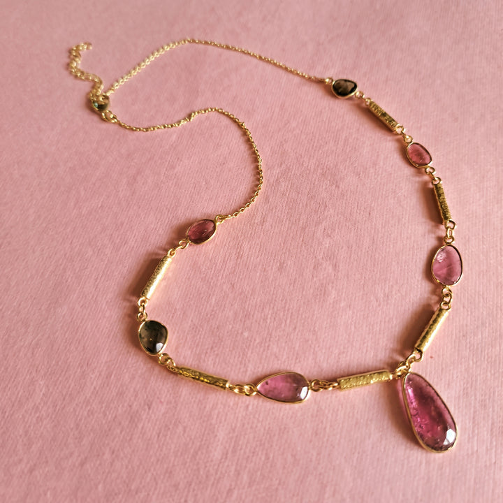 Gold necklace with green and pink tourmaline gemstones on a pink background.