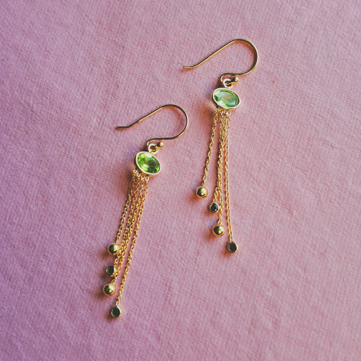 Green peridot hook earrings in gold featuring dangling garnet gemstones on a pink background.