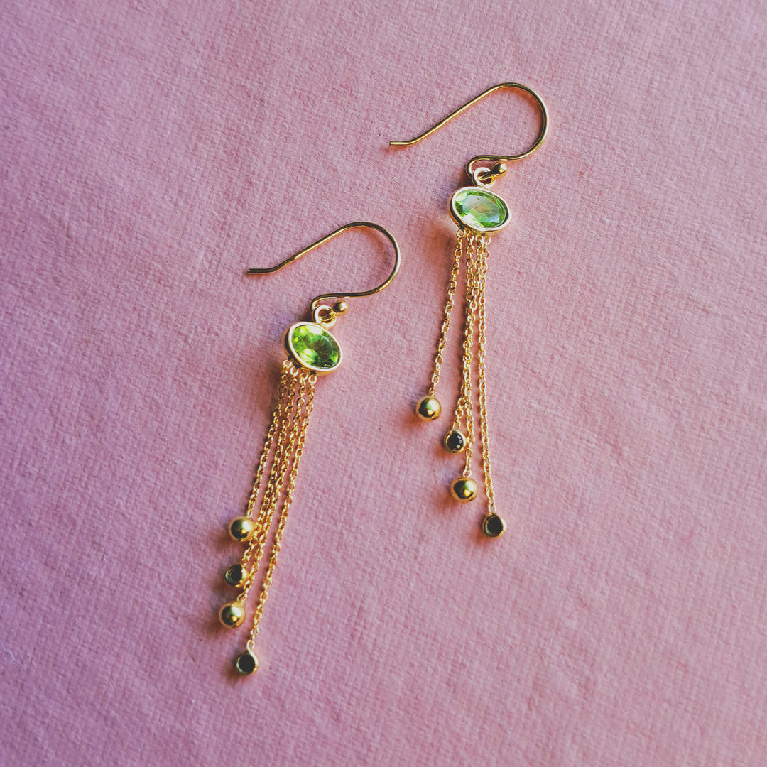 Green peridot hook earrings in gold featuring dangling garnet gemstones on a pink background.