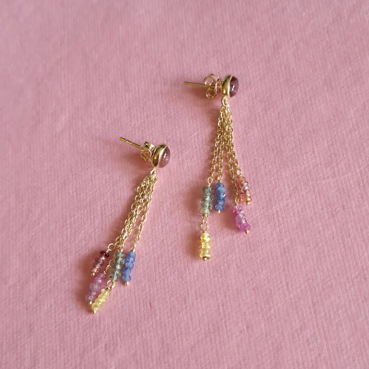 Dainty gold dangle earrings with Pink Tourmaline and blue  sapphire chain drops on a pink background.