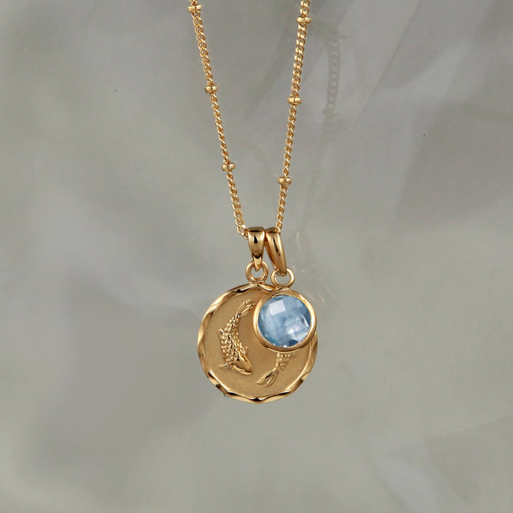 Gold zodiac necklace with pisces star sign and blue topaz birthstone charm on gray background
