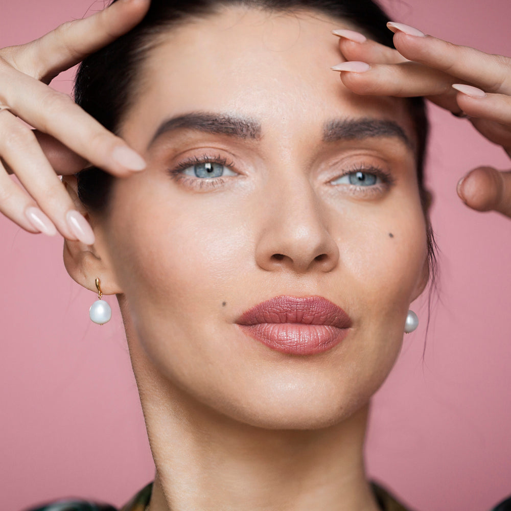 Close up of woman wearing gold and pearl stud earrings on a pink background