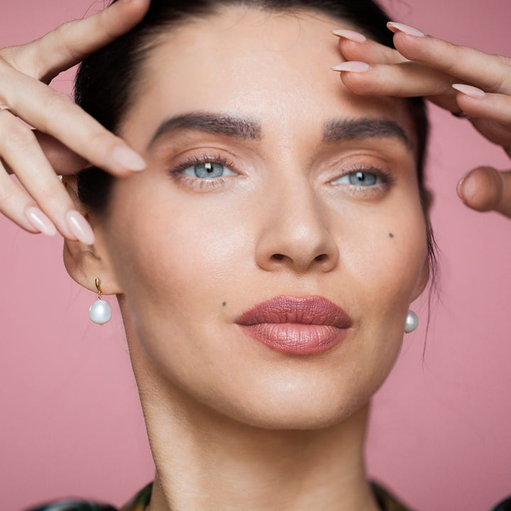 Close up of woman wearing gold and pearl stud earrings on a pink background