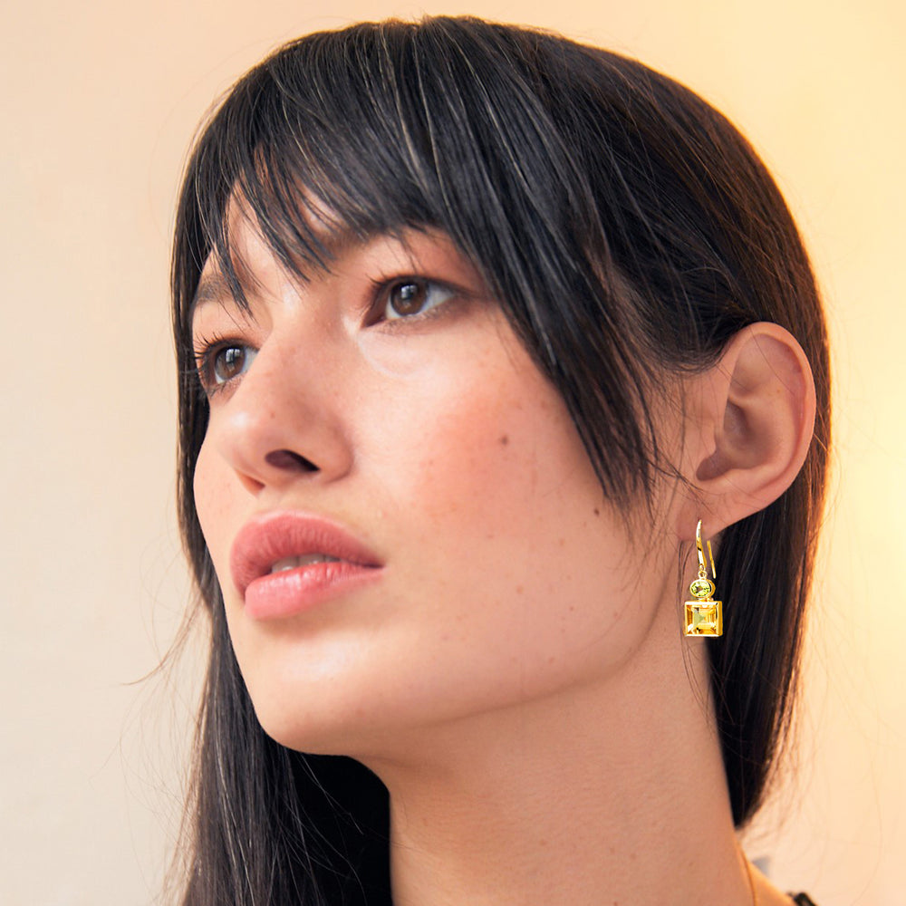 Close-up of a woman wearing square gold drop earrings with citrine and peridot gemstones