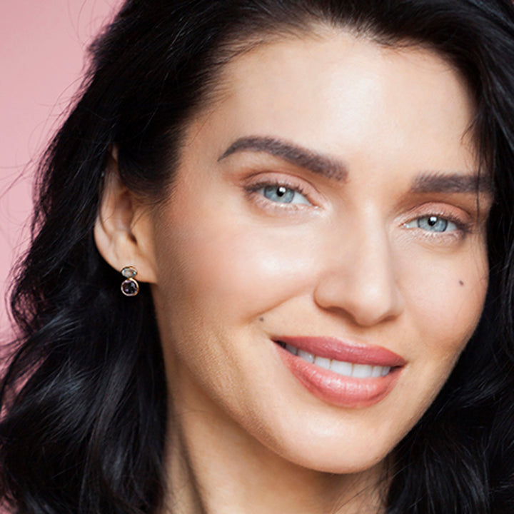 Close-up of a woman wearing gold stud earrings with amethyst and blue topaz gemstones against a pink background
