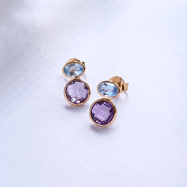 Pair of gold earrings with purple and blue gemstones on a light gray background
