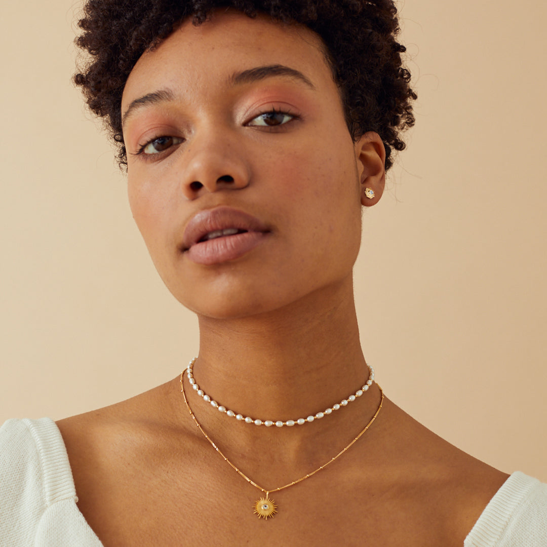 Model wearing a gold sun necklace and post earring set with white topaz gemstones and a pearl choker.