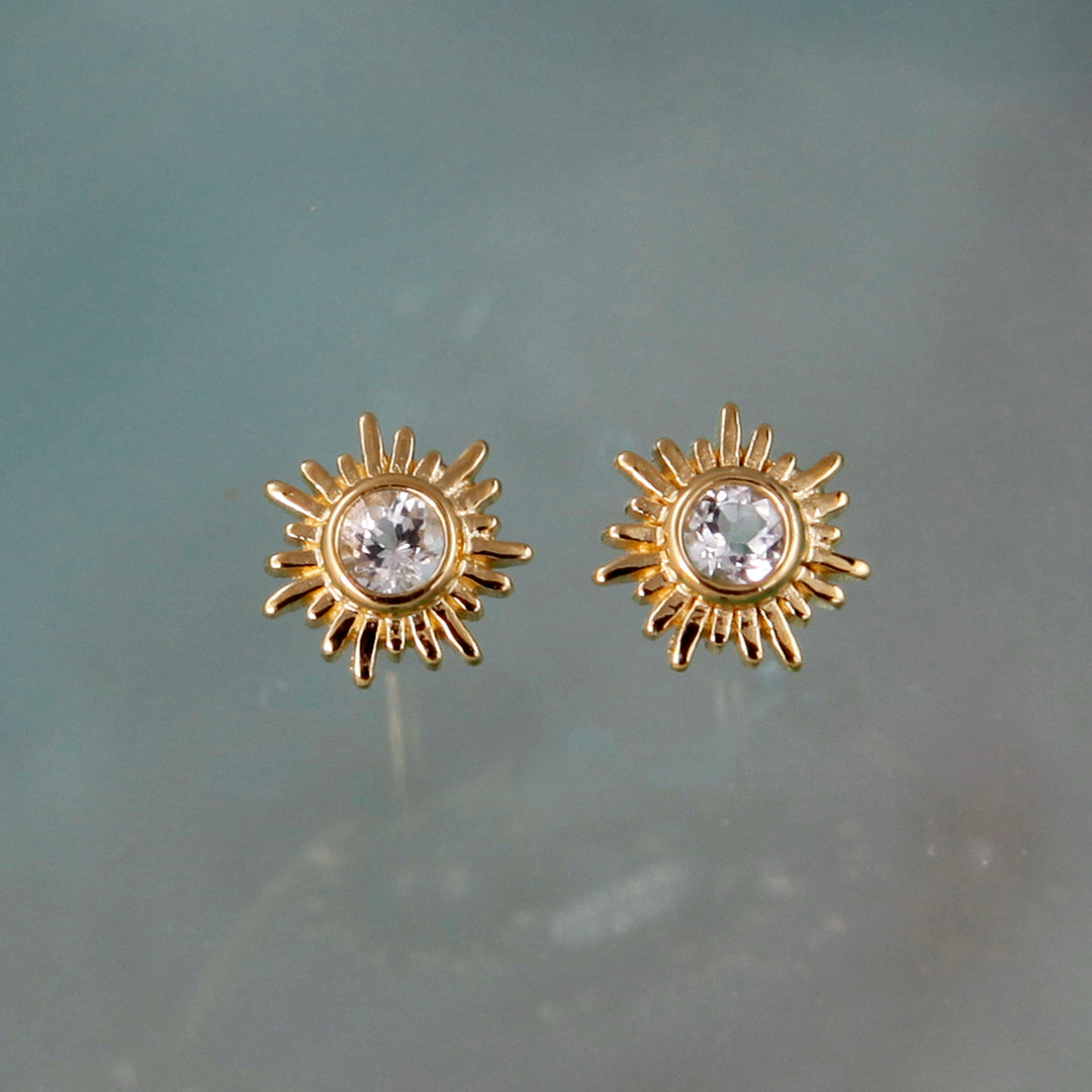 Gold sun stud earrings with white topaz centers on gray background