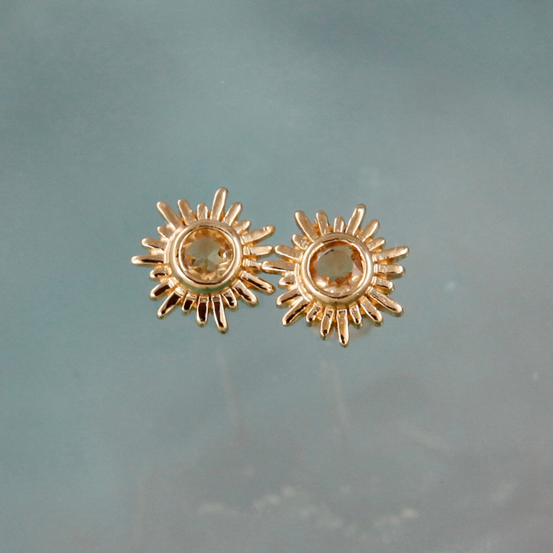 Gold sun stud earrings with citrine centers against an aqua background