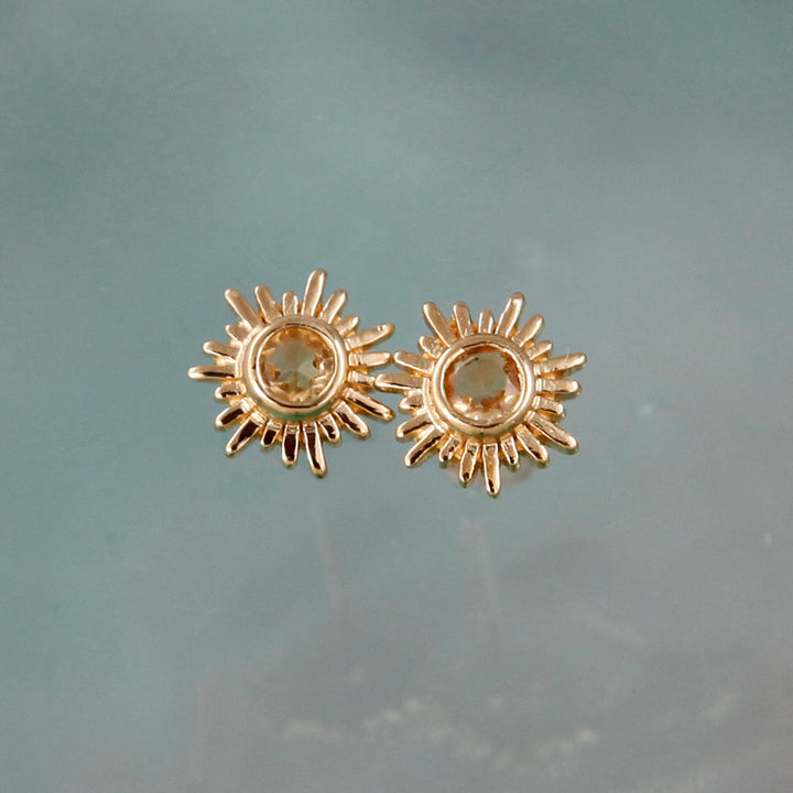 Gold sun stud earrings with citrine centers against an aqua background