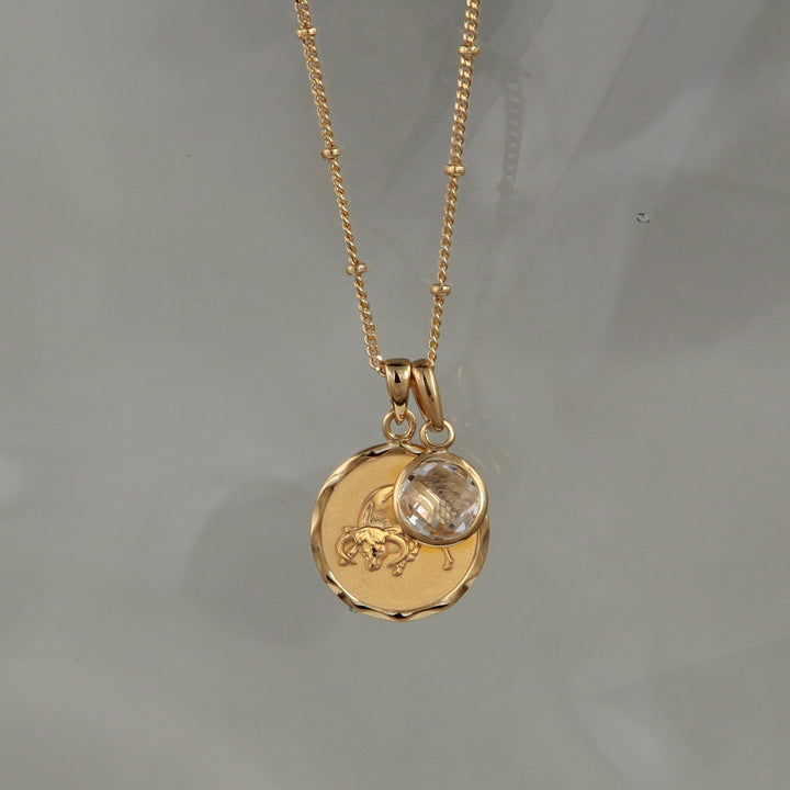 Gold Taurus star sign pendant necklace with a white topaz April birthstone charm on a gray background