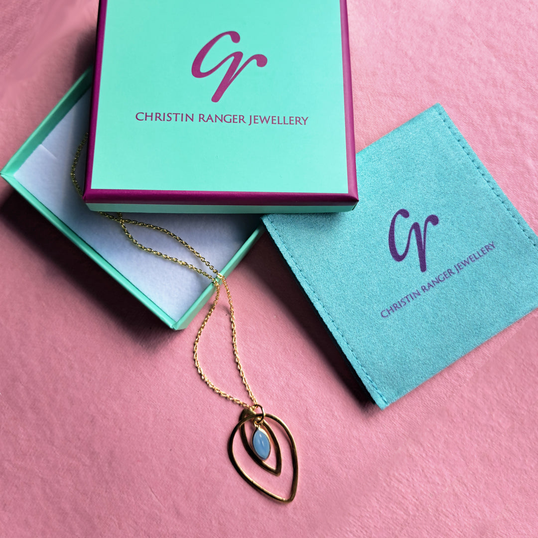 Christin Ranger Jewellery  gift packaging featuring a gold hoop necklace with a blue chalcedony gemstone on a pink background.