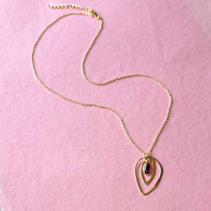 Alternative view of a gold teardrop shaped necklace with a garned gemstone on a pink background.