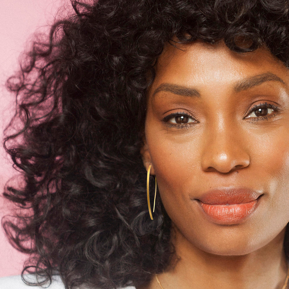 Woman with curly hair wearing a gold threader earring against a pink background