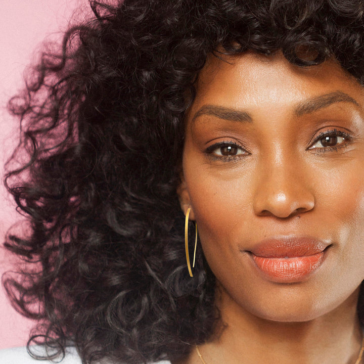 Woman with curly hair wearing a gold threader earring against a pink background