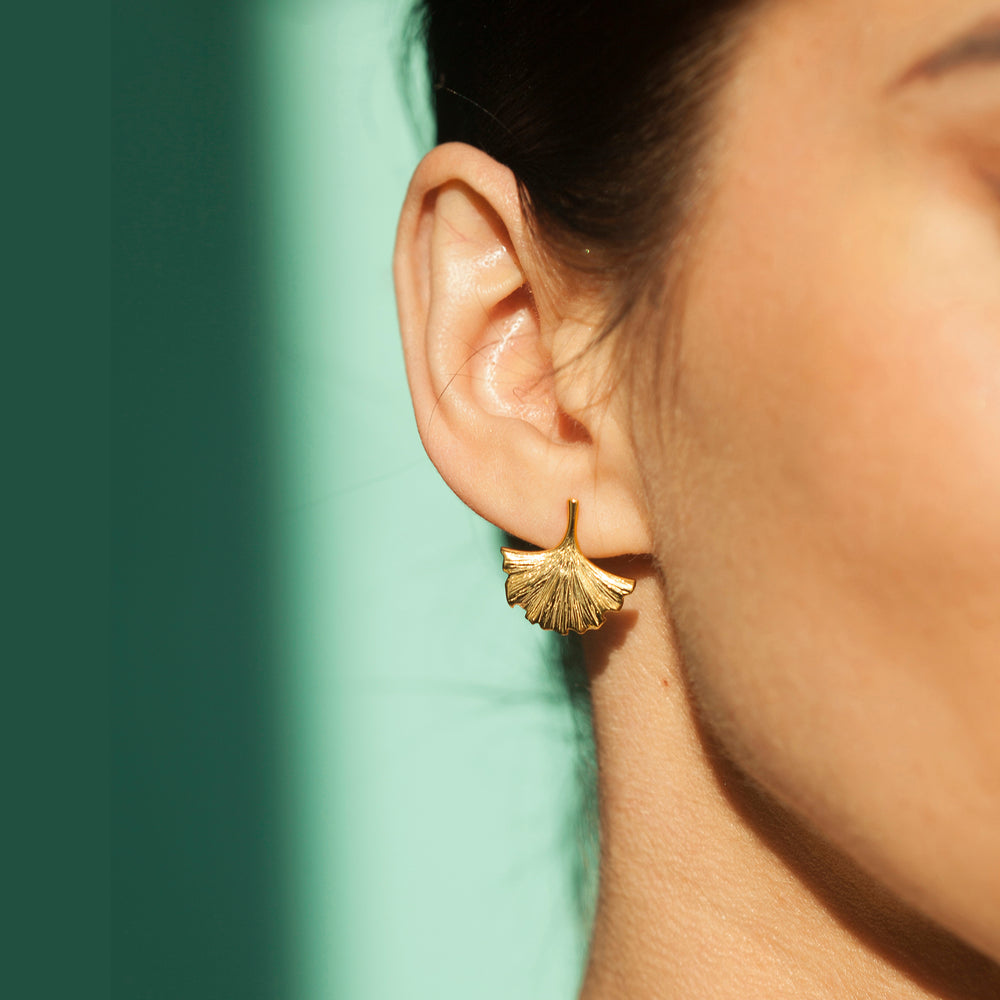 Close-up of a person wearing a gold ginkgo leaf earring against a green background